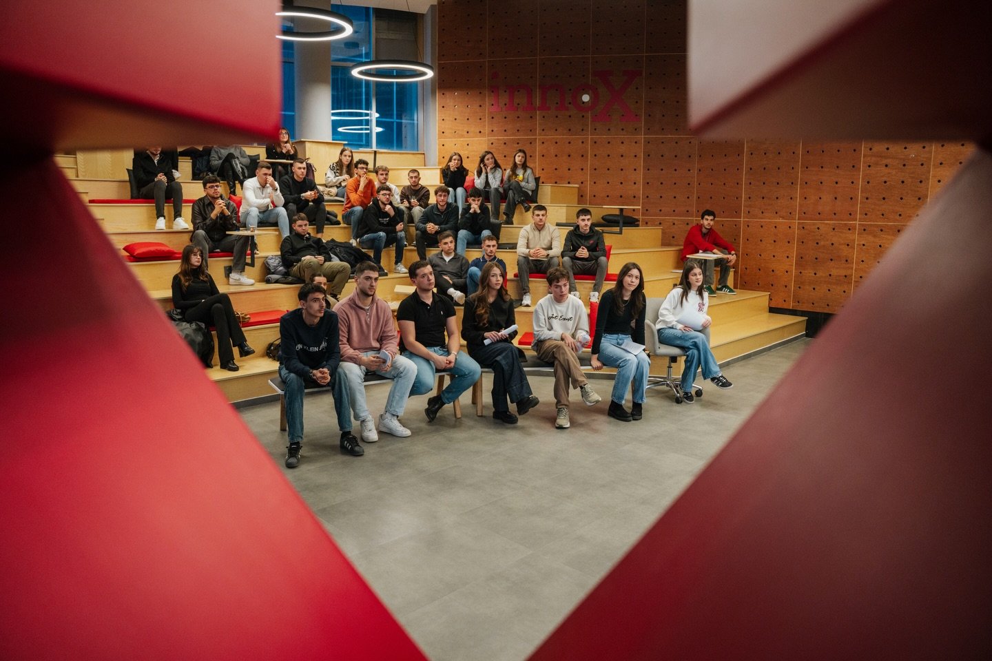 Young participants on the tiered seating at InnoX Center, framed by a red sculpture, during the From Idea to Business workshop